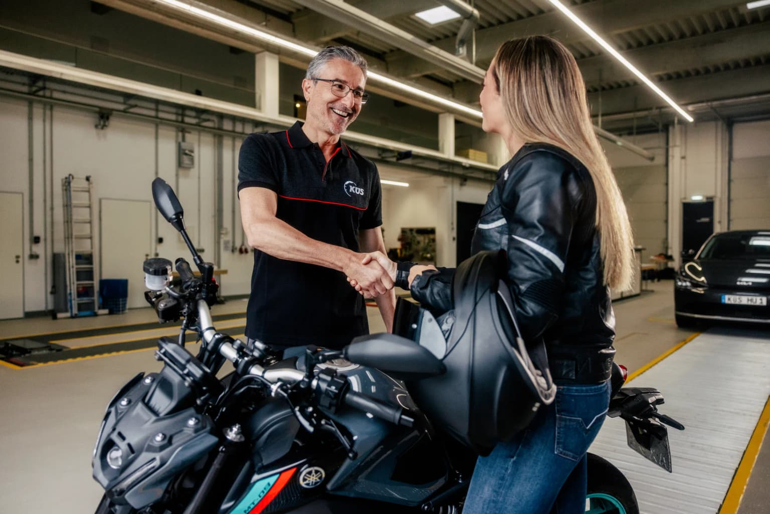 Hauptuntersuchung Motorrad in Köln-Mülheim KÜS-Prüfer begrüßt eine Kundin mit Helm am Motorrad zur Hauptuntersuchung in Köln-Mülheim
