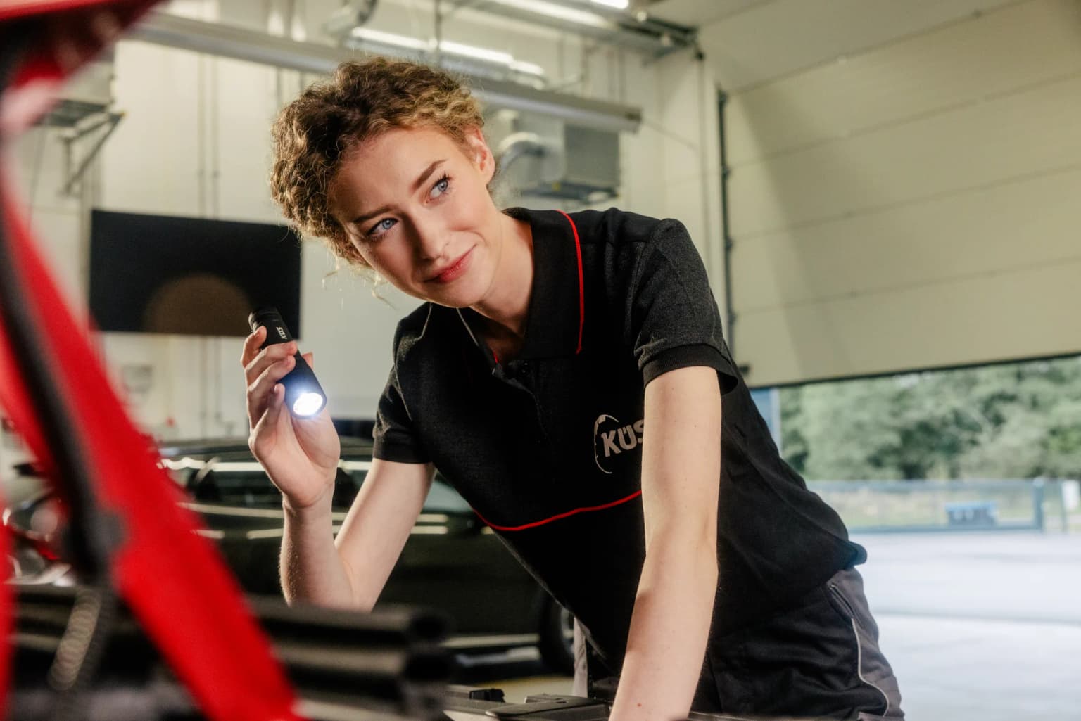 KÜS-Prüferin begutachtet mit der Taschenlampe den Motorraum eines Fahrzeugs